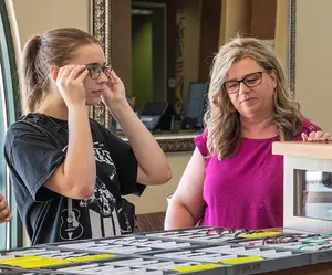 A woman is trying on glasses in front of a woman with blonde hair in a pink shirt at an optical shop