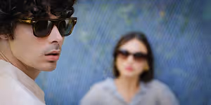A man with curly hair wearing sunglasses is looking at a woman wearing sunglasses in front of a blue wall