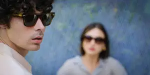 A man with curly hair wearing sunglasses is looking at a woman wearing sunglasses in front of a blue wall