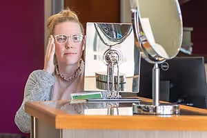 A woman sitting in front of a mirror wearing glasses and a necklace