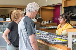 Three people are standing in front of a table with eyeglasses on it, with one of them wearing glasses and a necklace, and another one looking at the eyeglasses while the third one is looking at her.