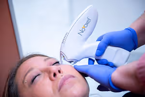 A person is using a Novoxel device on a woman's face, while the woman is sitting and closing her eyes.