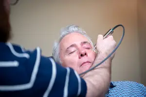 A man in a blue shirt is having his eye examined by a medical professional using an ophthalmoscope.