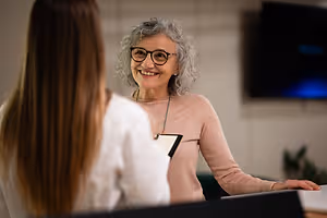 Woman with gray hair and glasses smiling at a young woman in a white shirt during a meeting in an office