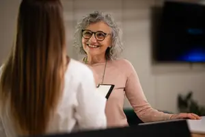 Woman with gray hair and glasses smiling at a young woman in a white shirt during a meeting in an office