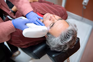 An elderly man is undergoing a laser treatment for skin rejuvenation while wearing blue gloves and a maroon shirt.