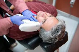 An elderly man is undergoing a laser treatment for skin rejuvenation while wearing blue gloves and a maroon shirt.