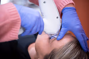 A woman is getting her nose pierced with a piercing gun while lying on a bed with her head down and a person wearing a pink sweater is holding the piercing gun and wearing blue gloves.
