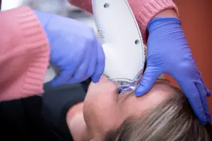 A woman is getting her nose pierced with a piercing gun while lying on a bed with her head down and a person wearing a pink sweater is holding the piercing gun and wearing blue gloves.