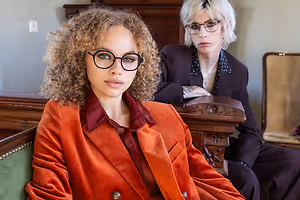 Two women sitting next to each other on a chair, one wearing glasses and a necklace, and the other wearing a blazer with a polka dot shirt, sitting in a room with wooden furniture and a white wall