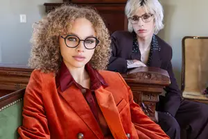 Two women sitting next to each other on a chair, one wearing glasses and a necklace, and the other wearing a blazer with a polka dot shirt, sitting in a room with wooden furniture and a white wall