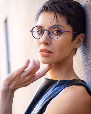 A woman with short hair wearing glasses and a black sleeveless top is posing for a photo while touching her chin.