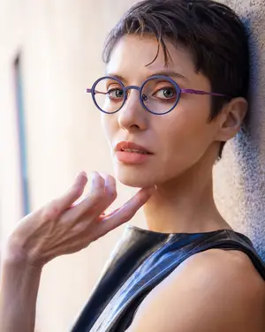 A woman with short hair wearing glasses and a black sleeveless top is posing for a photo while touching her chin.