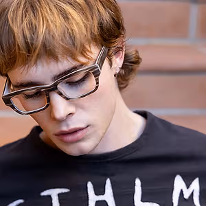 A young man wearing glasses is looking downward. He has short brown hair and is wearing a black t-shirt with white writing on it.