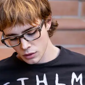 A young man wearing glasses is looking downward. He has short brown hair and is wearing a black t-shirt with white writing on it.