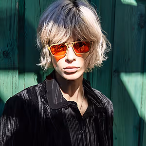 A man wearing a black shirt and sunglasses with reflective lenses poses against a green wooden wall.