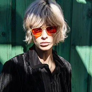 A man wearing a black shirt and sunglasses with reflective lenses poses against a green wooden wall.
