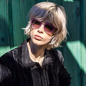 Blonde woman wearing purple sunglasses and a black shirt against a green wall
