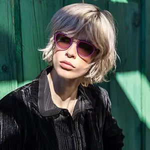 Blonde woman wearing purple sunglasses and a black shirt against a green wall