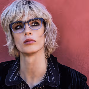 A woman with short blonde hair and glasses poses for a photo against a red wall