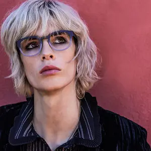A woman with short blonde hair and glasses poses for a photo against a red wall