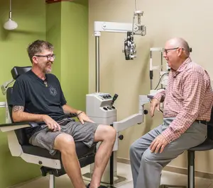 Two men are sitting in a room with medical equipment in the background, one man is wearing glasses and the other man is wearing a plaid shirt and a belt.