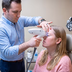 A man in a blue shirt and a woman in a pink sweater are in a medical setting where the man is performing tixel i treatment on the woman's eye.