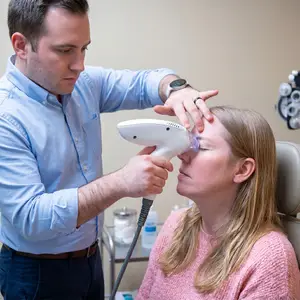 A man in a blue shirt and a woman in a pink sweater are in a medical setting where the man is performing tixel i treatment on the woman's eye.