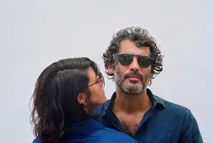 A man with curly hair and a beard wearing sunglasses and a blue polo shirt is standing next to a woman wearing glasses and a blue long-sleeved shirt, both are standing in front of a white wall and seem to be looking at something.