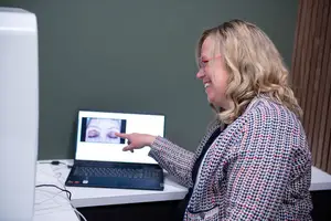 A smiling woman with glasses points at a laptop screen showing a close-up of eyes