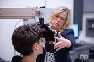 A woman optometrist examines a young boy's eye using specialized equipment in an office setting.