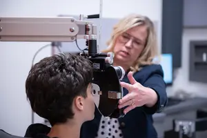 A woman optometrist examines a young boy's eye using specialized equipment in an office setting.