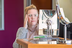 A woman with glasses is looking at her reflection in a mirror with a reflection of another mirror and a wooden desk with a silver stand and a monitor with a reflection of a person and a wall in the background.