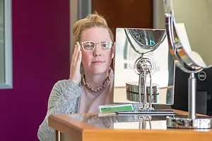 A woman with glasses is looking at her reflection in a mirror with a reflection of another mirror and a wooden desk with a silver stand and a monitor with a reflection of a person and a wall in the background.