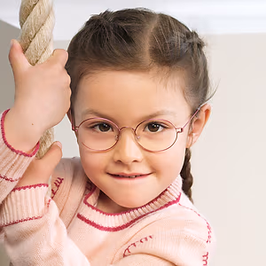 A little girl wearing glasses and a pink sweater is smiling and holding onto a rope.