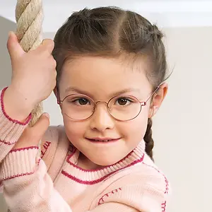 A little girl wearing glasses and a pink sweater is smiling and holding onto a rope.