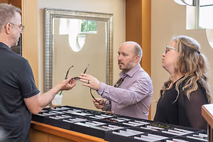 Three adults are standing in front of a display case of glasses and one of them is holding a pair of glasses.