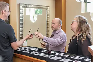 Three adults are standing in front of a display case of glasses and one of them is holding a pair of glasses.