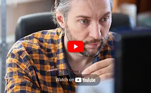 A man wearing glasses and a plaid shirt is looking at a computer monitor with a YouTube logo on it