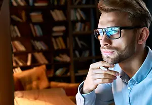 A man wearing sunglasses and a light blue shirt is sitting in a chair in a dimly lit room with bookshelves and a couch in the background.