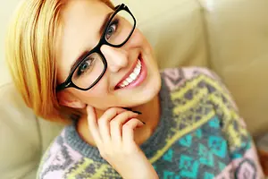 Portrait of a woman sitting on a couch wearing glasses and a colorful sweater, with a pencil in her hand and a smiling expression