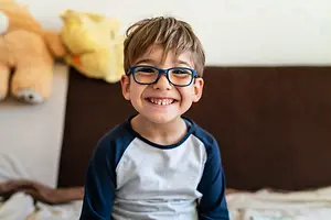 A young boy wearing glasses is smiling on a bed with stuffed toys in the background