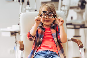 A girl in a red shirt and jeans sits in a white chair in an optometrist's office, wearing black ophthalmoscopes on her eyes and smiling happily.