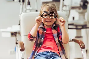 A girl in a red shirt and jeans sits in a white chair in an optometrist's office, wearing black ophthalmoscopes on her eyes and smiling happily.