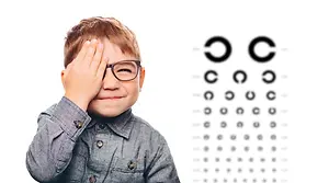 A young boy wearing glasses and covering one eye while smiling, standing in front of an eye chart.