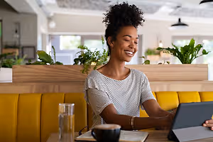 A woman sitting at a table in a restaurant booth, smiling while using a tablet.