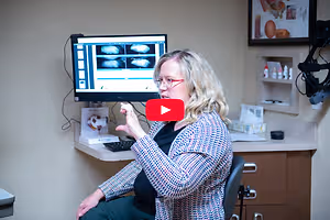Optometrist in an examination room gesturing beside a monitor displaying eye images
