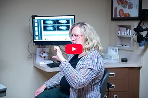 Optometrist in an examination room gesturing beside a monitor displaying eye images