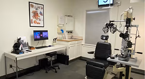 A room with an eye examination chair and a desk with a computer monitor, a stool, a cabinet, and a monitor on the wall.