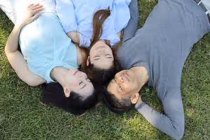 Three people laying on a grass field in a park with their eyes closed and smiling